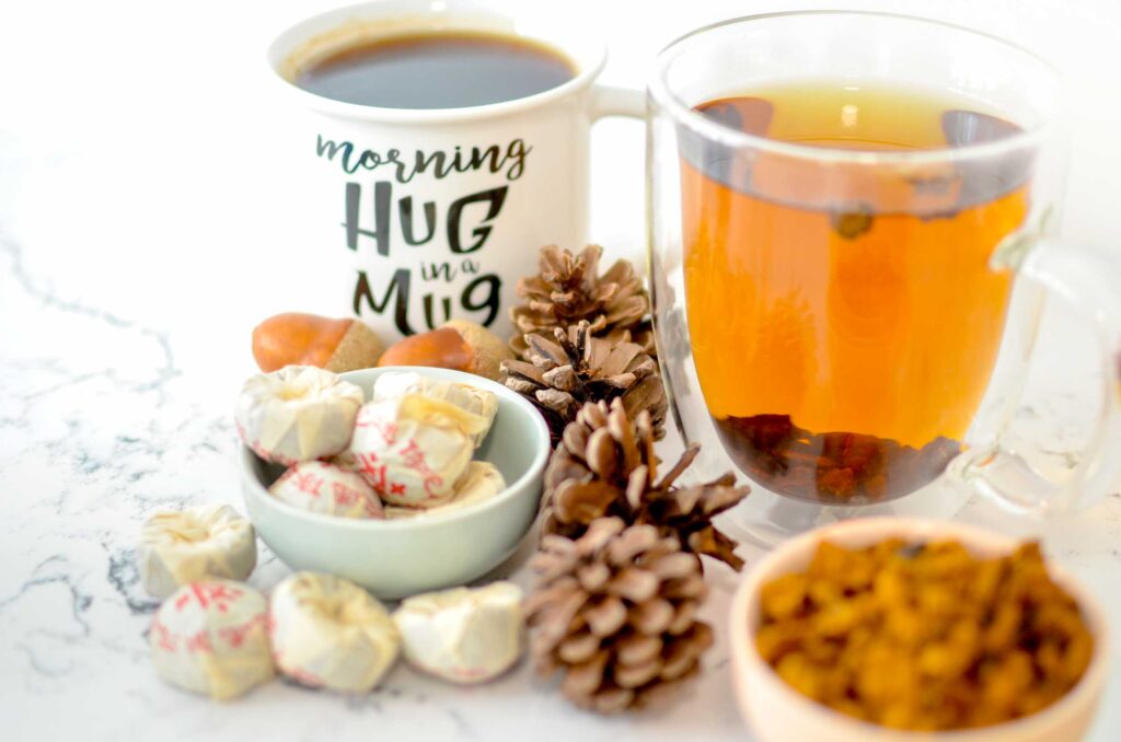 two mugs. One is filled with a dark brown brew, there are pu'erh tea cakes in front of it. The other is a double walled mug with orange brew, there are pieces of chaga mushroom tea in a pinch bowl in front. 
