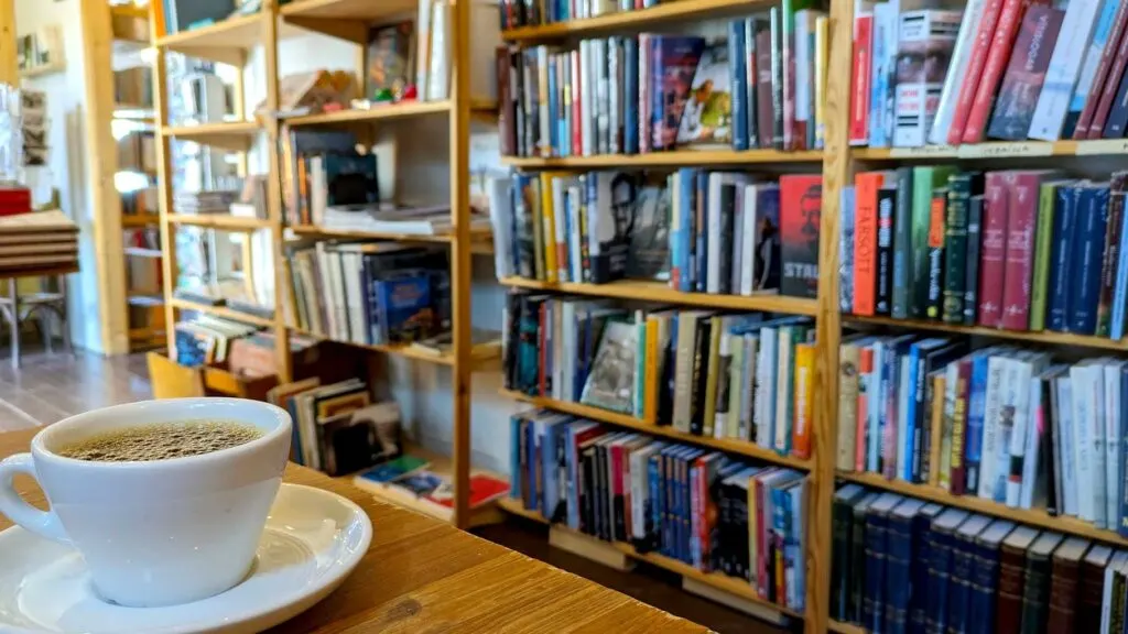 a white mug of black coffee, still very hot, sitting on a table. Behind the coffee are slightly out of focus bookshelves filled with books