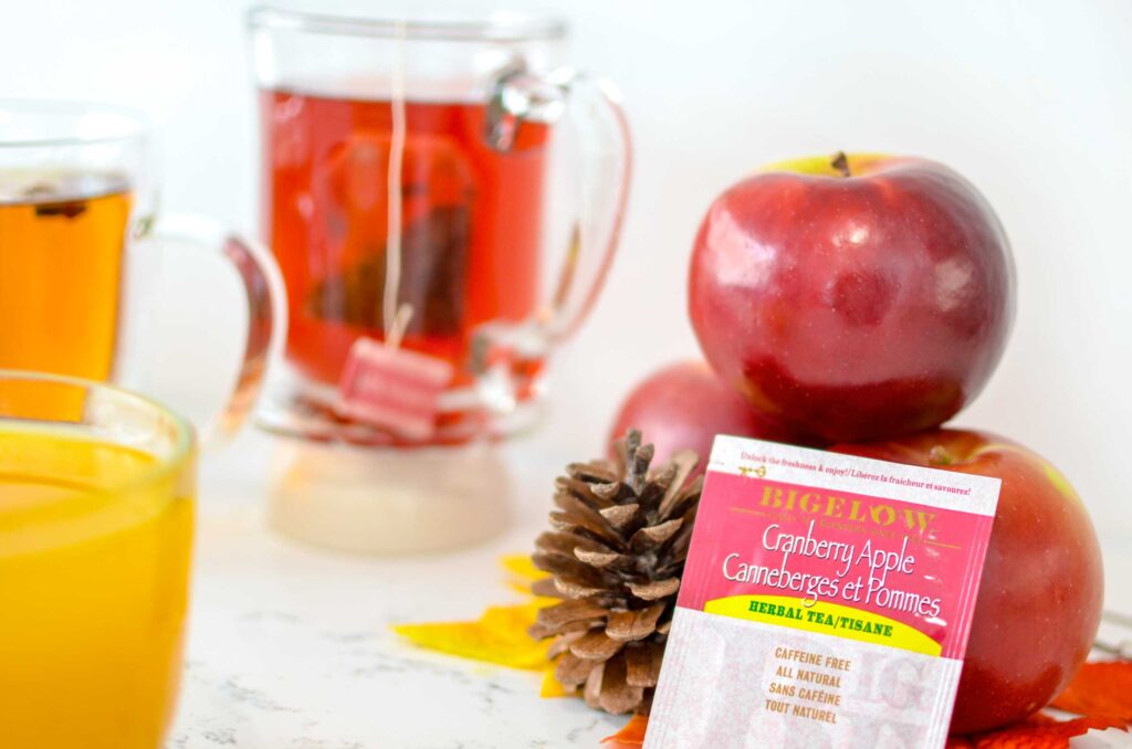 cranberry apple individual tea bag for fall is resting on a tower of shiny macintosh apples and a pinecone. Behind it is a clear glass mug with a tea bag floating inside, it's a red colour and the tea bag string is hanging outside. But it's too out of focus to see what it says. On the left, you can also see two glass mugs filled with orange and yellow tea. 