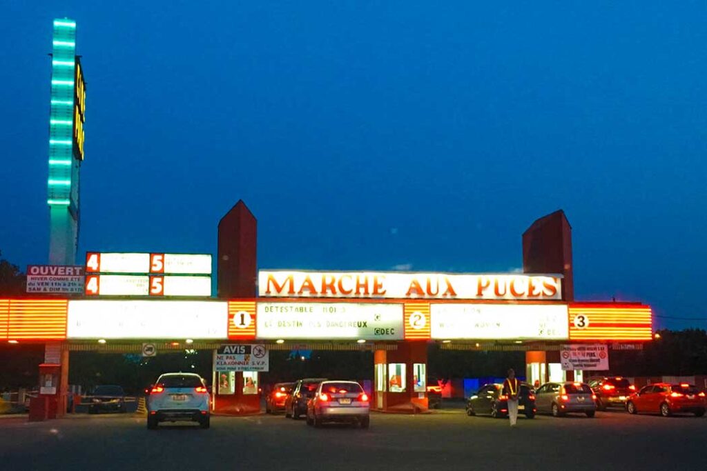 drive in montreal at dusk. This is the entrance to the drive in, cars are lined up under a lit up sign that says: Marche aux Puces. You can almost make out the times and movie showings above.