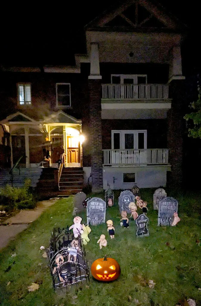 a haunted decorated house in montreal for halloween at night. A vertical image shows a lawn filled with gravestones, creepy standing baby dolls with black marker drawn on their face , a gated off cage with more dolls trapped inside. There's also a carved pumpkin with light shining through it's face.