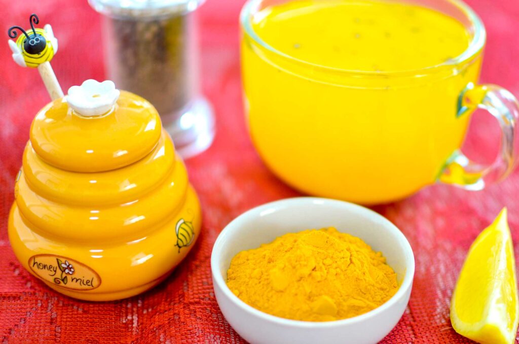 a close up of a honey jar, it looks like a beehive with a little bee on a stick sticking out of the cover. There's a painted bee on the jar, and it says honey miel. Next to it is a pinch bowl filled with turmeric powder, a pepper grinder, and a mug of yellow turmeric tea, and a lemon wedge on the corner