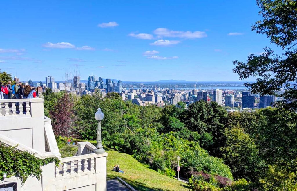montreal summit circle lookout. There's a stone railing and stairs leading up to it. It overlooks a green hill with trees below and in the background are the buildings of Montreal city skyline. Behind it you can even see the st. lawerence river and some mountains on a completely clear day