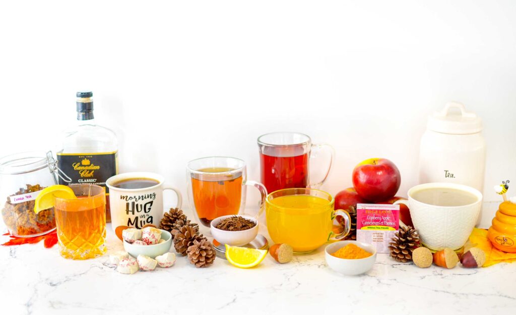 all the non spiced fall teas sitting on a marble counter with a white wall behind.

From the left: a jar of dried chaga tea, Canadian club whiskey, with a tumbler of amber liquid in front. A mug with a dark brown brew, the mug says morning hug in a mug. In front sit a pinch bowl with wrapped pu'erh tea cakes.
Pinecones separate the brewed orange tea, with a pinch bowl of dried hojicha tea leaves and a lemon wedge in front. Then there are two mugs, one with a red brew, the other with a golden yellow. In front of the yellow brew is a pinch bowl filled with yellow turmeric powder. Then there's a tower of three apples, with a tea bag package for Bigelows Cranberry apple tea sitting on it. And finally you have a white cappuccino mug with a light brown latte. There's a tea jar behind it, and a honey jar cut off as the photo ends on the right