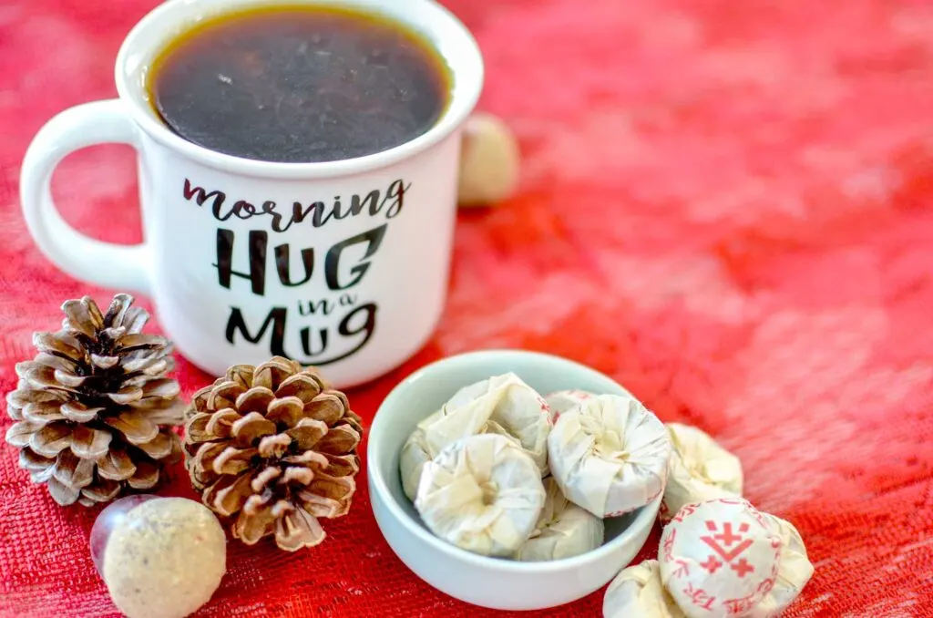 a white mug with the text: morning hug in a mug written on the front. It's filled with a dark brown brew: puerh. Next to it is a punch bowl, it's filled with individually wrapped pucks, or tea cakes. there's Chinese writing on it. The background is a red table cloth, with scattered pinecones and acorns around the subjects. 