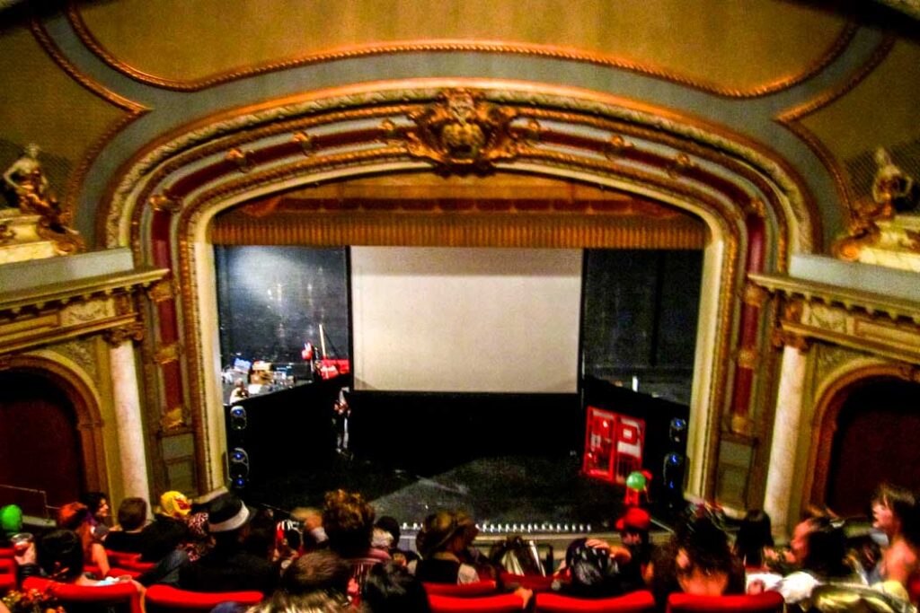 The theater at the rocky horror picture show in montreal on halloween before the show starts. The seats are int he mezzanine, the chairs are red and already filled with people dressed in various costumes. You can see the stage below with a projector screen in the middle (to show the movie at the same time). The theatre itself is grand with light blue and gold decor.  