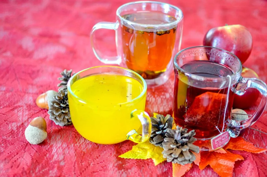 three teas in different glass mugs that arent spiced for fall. They're on a bright red table cloth with pinecones and acorns, and fall coloured leaves around them. 

The glass mugs let you see the beautiful bright colours of the tees: a bright yellow turmeric tea, an orange chaga tea, and a deep red cranberry apple tea