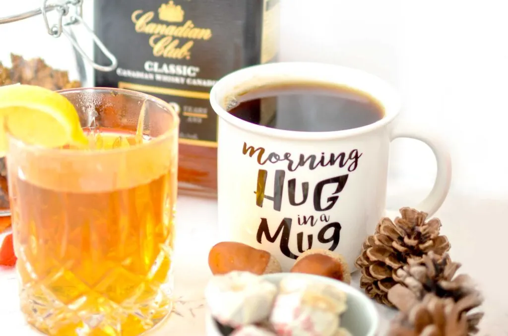 two glasses are sitting on the counter. On the left is a whisky tumbler, filled with an amber drink, and a lemon slice on the rim. Next to it is a white ceramic mug, filled with a dark brown brew. In the back, out of focus, is Canadian Club classic whiskey bottle.