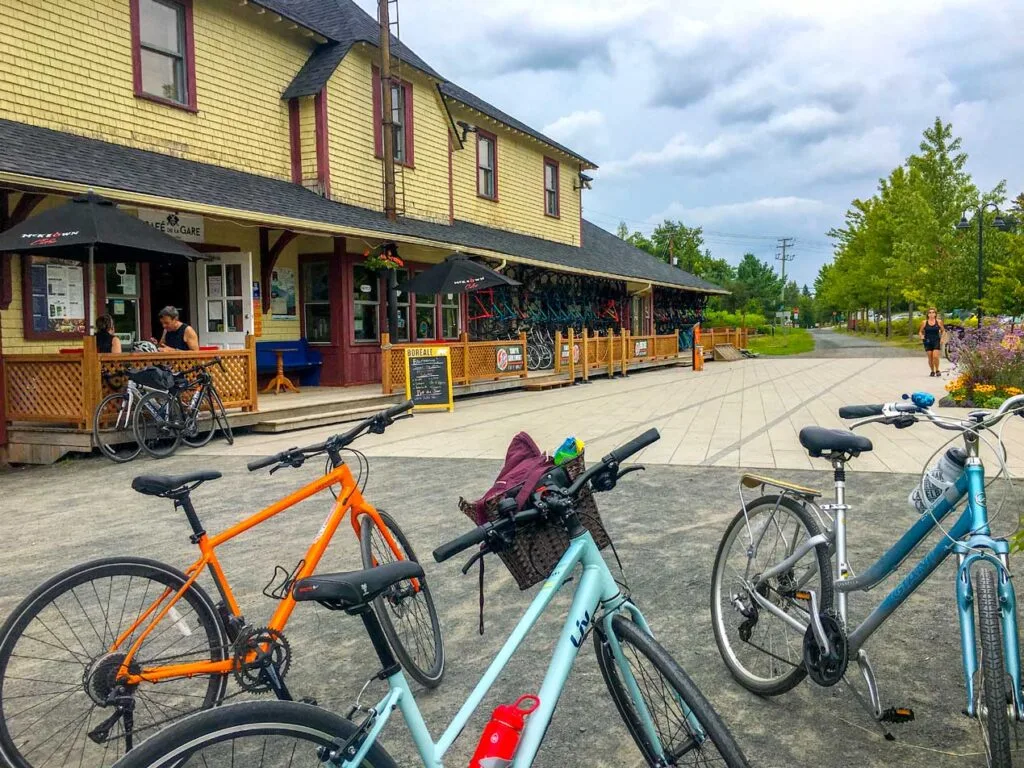 biking ptit train du nord st sauveur. three bikes are leaning on their stands on the bottom third of the image. Two are blue, and one is a bright orange. On the left going all the way to the middle of the photo is an old train station building. Today, it has a cafe with seats outside, and a bike rental shop with bikes all lined up outside. The path continues into nature.