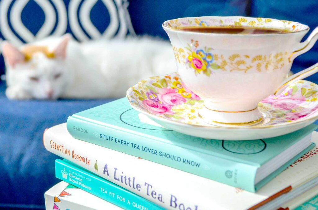 A stack of books, all seem to be matching with teal or white binding. On top sits a beautiful white and pink floral tea cup with piping hot tea. You can still see the steam rising from it. It's a cozy picture. Books, tea and there's even a cat sleeping in the background. He's matching the aesthetic of the image as he's white with orange as an accent colour around his ears. He's sleeping on a navy blue couch, and a blue and navy patterned piillow. It adds a nice deep rich grounding colour to the background of the scene.