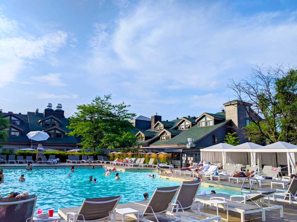 manoir pool in saint sauveur water park. Lounge pool side chairs are lined up on the patio in front of a large pool with adults and kids, but it still isn't too packed. There are cabanas on the right side. Behind the pool is the manoir saint sauveur. You can only see the large green roof with dormers sticking out from it. 