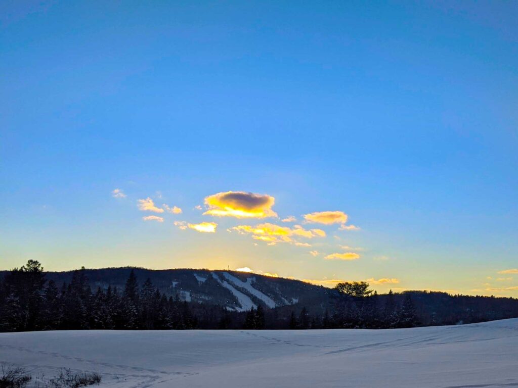 sauveur ski hill in the winter at sunset in the distance. A snow covered field takes up the bottom third of the image. The middle third is the ski hill, it doesn't look like much, just the shadow of a mountain, but there are white lines that go from the top to the bottom - these are the ski hills. The sun is setting behind the mountain, it leaves a yellow glow. You can count the number of clouds in the sky, but they are also a golden yellow, popping out against the blue sky. The sky is the rest of the image