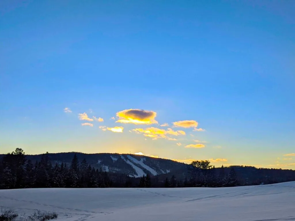 sauveur ski hill in the winter at sunset in the distance. A snow covered field takes up the bottom third of the image. The middle third is the ski hill, it doesn't look like much, just the shadow of a mountain, but there are white lines that go from the top to the bottom - these are the ski hills. The sun is setting behind the mountain, it leaves a yellow glow. You can count the number of clouds in the sky, but they are also a golden yellow, popping out against the blue sky. The sky is the rest of the image