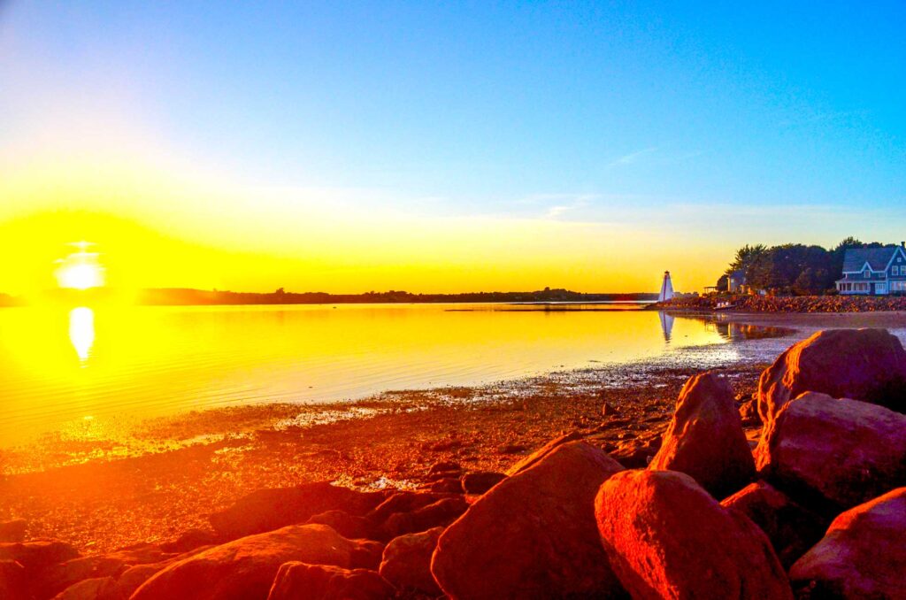 brighton beach lighthouse pei canada at sunset. The sun beams as it hits the horizon. Where the sun should be is a white spot, but around it emanates a large golden yellow glos, that spread across the horizon and into the water as a near perfect reflection. 

The glow leads you're eye to the shoreline, where the red rocks look even brighter. 

As you follow the shorelines down the beach, you reach a house and a white lighthouse with the perfect reflection in the water. 

