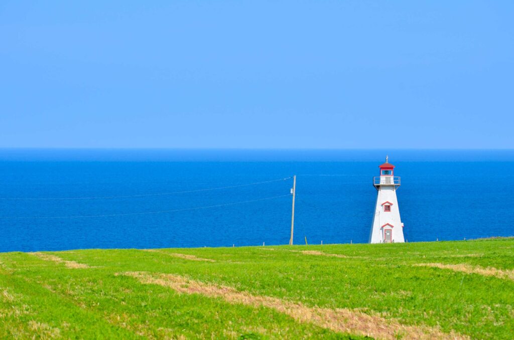 incredible stark view of the cape  tryon lighhouse in pei canada. 

The photo is separate into thirds horizontally, represented by vibrant green grass at the base, a deep blue for the sea in the middle, and a lighter shade of baby blue for the sky - without a cloud in sight.

On the right third of the frame stands a tall white structure with a red roof, small balcony at the top, a window, and a door. It starts at the base of the grass and reach almost to where the sky starts! 