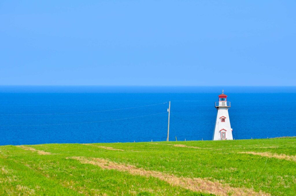 incredible stark view of the cape tryon lighhouse in pei canada. The photo is separate into thirds horizontally, represented by vibrant green grass at the base, a deep blue for the sea in the middle, and a lighter shade of baby blue for the sky - without a cloud in sight. On the right third of the frame stands a tall white structure with a red roof, small balcony at the top, a window, and a door. It starts at the base of the grass and reach almost to where the sky starts!