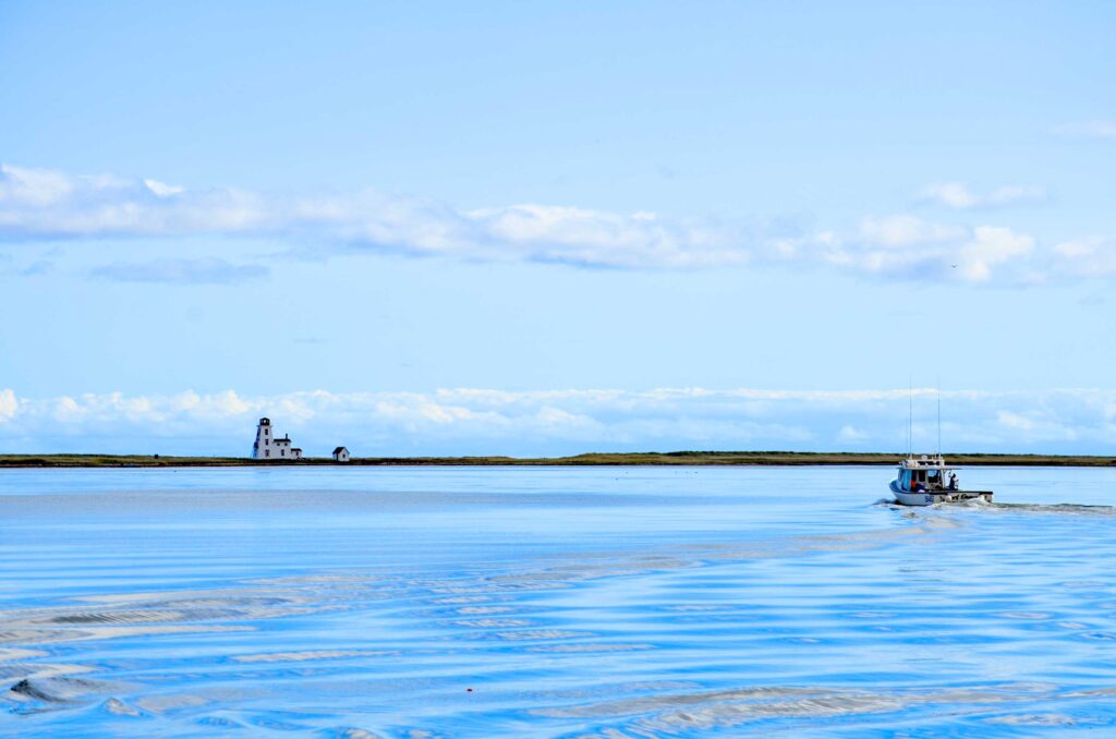 cascumpec floating lighthouses prince edward island. 
The photo is cut in half horizontally by a narrow sand dune going the entire way across. Above the sand dune is the sky with a thin layer of clouds. Below it, the water. 

A third of the way on the sand dune is a three story light house and home. On the right side, you notice ripples in the water that lead you to a small boat making it's way to the lighthouse