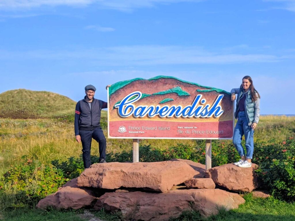 a man standing on red rocks with his arm on a large sign that reads Cavendish, Prince Edward Island National Park.  On the right side of the sign, is a woman leaning on the sign and also standing on the same bed of red rocks. 

Around the red rocks is a field of large lush green grass, and far in the distance, you can see a sand dune covered in grass. 