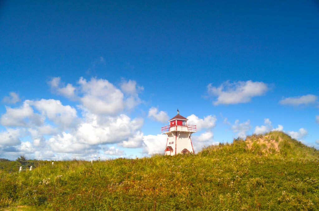 covehead lighthouse pei popping up behind the messy grass growing on sand dunes. It's white and red, and not the tallest of the pei lighthouses. The clouds aer in perfect puffs, but only across the middle of the photo, it creates the appearance that they are moving. 