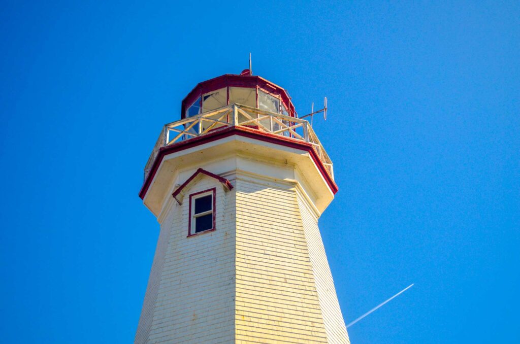 close up of the top of the east point lighthouse on prince edward island. You can see the battered shingles, and the barrier at the top. 

The sky is a bright blue, and empty besides for the streak of a flying plane behind the lighthouse