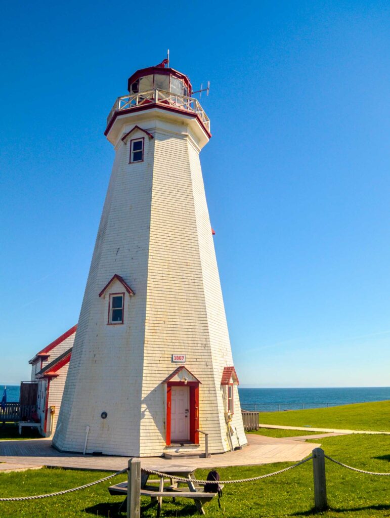 Vertical image: east point lighthouse on-the edge of pei. There's a nice wood walkway leading and surrounding the base of the lighthouse. The lighthouse looksl ike it could be painted white again, it's looks a bit brown in some areas. But the red accent paint around the windows, doors, and around the top still looks vibrant. Once the grass ends, you see the water all around it. What you don't see is the drop off cliff!