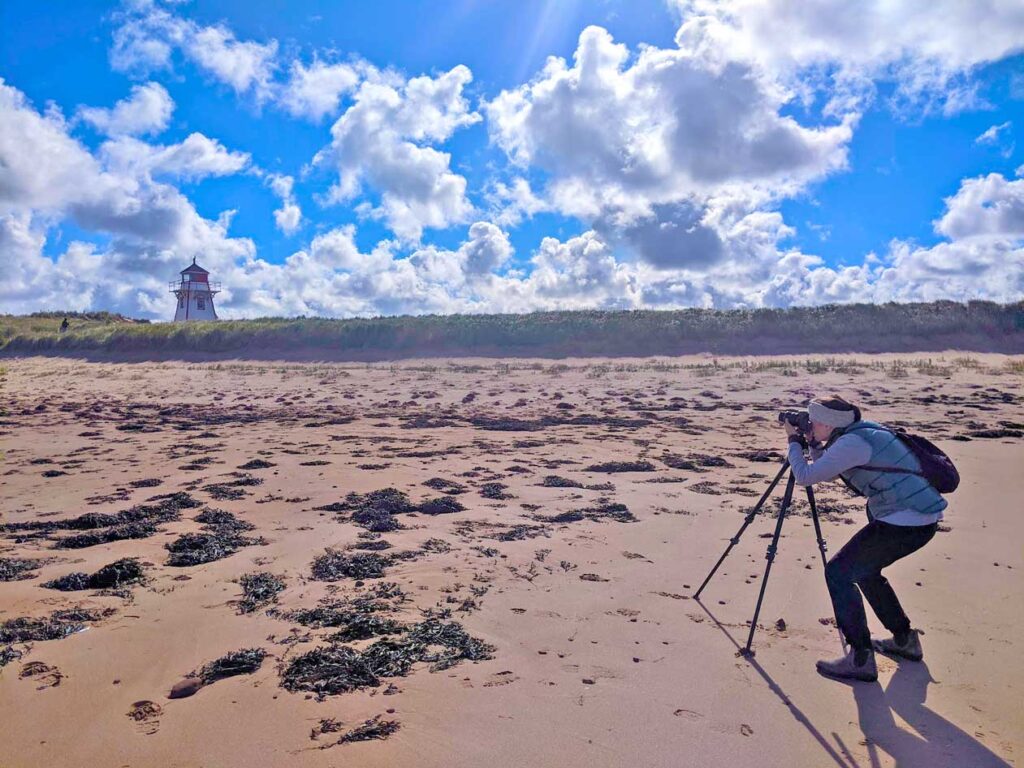 a woman in the right of the photo standing behind a camera on a tripod. She's on the great expanse of the beach, photographing the covehead lighthouse in the distance. 