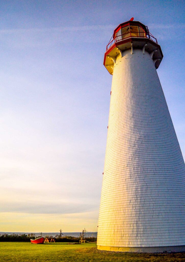 point prim first prince edward island lighthouse at golden hour. 

A vertical image to show the heigh of the point prim lighthouse towering above the land. 

The grass as a yellow hue, as the sun sets, it also shines on the left side of the lighthouse. The back of the lighthouse is more in shadows. 

The sky is very relaxing, with whispy clouds turning the sky into calming yellow and blue hues. 