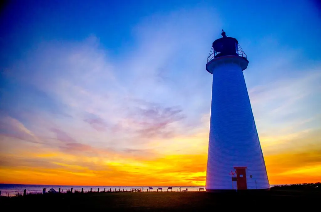 points prim lighthouses of prince-edward island canada at sunset. 

It's a wide angle view. The land is almost completely in shadows. The lighthouse stands tall way up into the sky, it has a blueish gradient getting darker the higher up. 

Behind the lighthouse the sunset is radiant. The sun is about to meet the horizon, creating a glowing orange and yellow hue across the bottom. There's good cloud covering near the horizon and it clears up towards the top of the image as the sunset turns from yellows, to purple, to back to blue. 