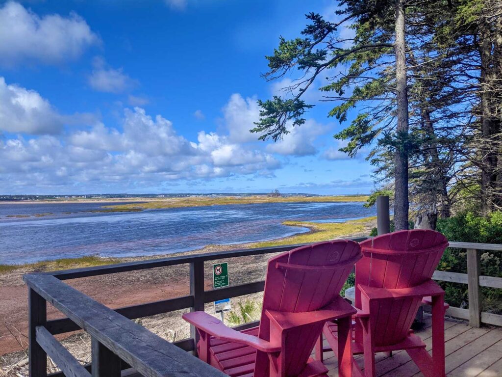 two red chairs sit on a raised wood balcony overlooking the prince edward island national-park.The red sand turns into grass before it meets the water and eventually meets the sky all the way in the distance. 