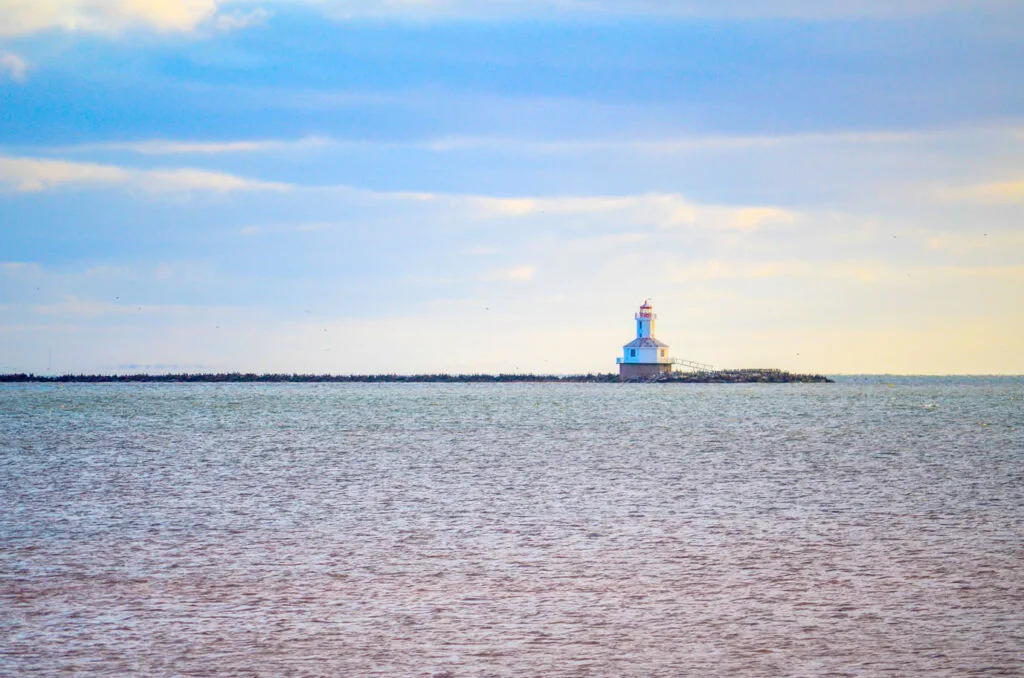 a very narrow inlet of land cuts the photo in half, separating the sky and the water. Three quarters of the way across the land, sit a tiny lighthouse on a slab of concrete: the indian head lighthouse pei canada. 

The water towards the bottom of the photo actually has a reddish tint to it, this is how you know you're in prince edward isalnd. 

The sun must be setting soon, because the sky also has a calming yellow tint to the clouds and close to the horizon. 