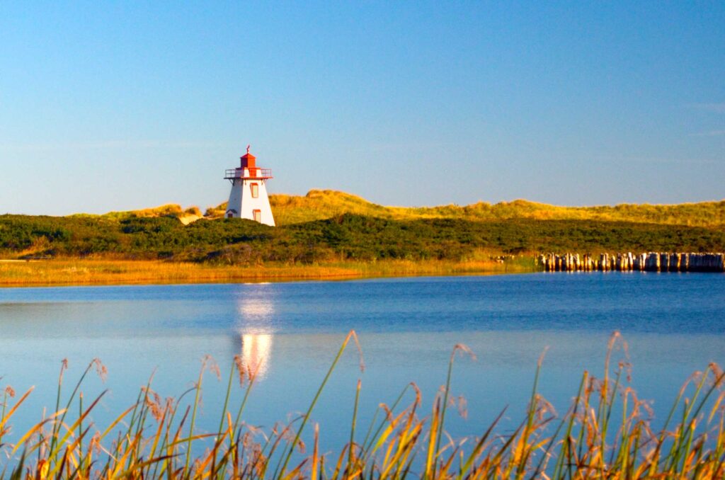 st peters harbour lighthouse pei.  There are wheat grass sticking out from the bottom of the photo, framing the entire edge. Then you reach the calm blue water of the river, you can see a blurred white reflection of a tall structure. When your eyes meet the land, it's of deep green hill, and a bright white and red lighthouse protruding from the landscape reaching the clear blue sky above. 