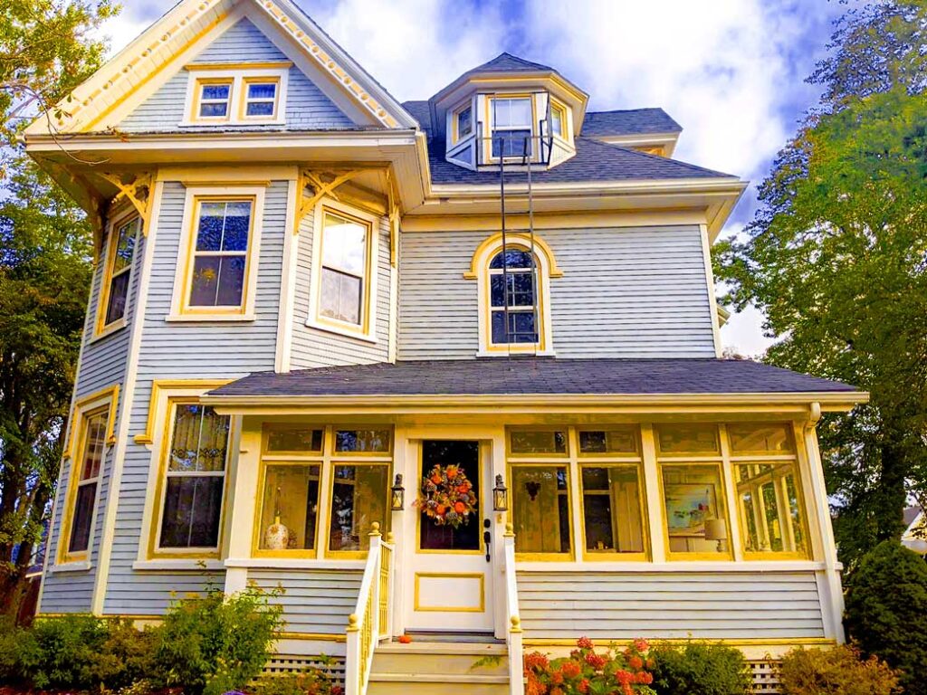 summerside hotels on prince edward island. A large victorian style home takes up 90% of the image. It has light blue patined shingles, with bright yellow accents around the doors and windows. It's a three story house, starting with an invited windowed solarium in the front of the house. There's a protruding tower on left side, offering bay windows on the first two stories of the house. Finally the last floor is the roof with a small dormer sticking out. 