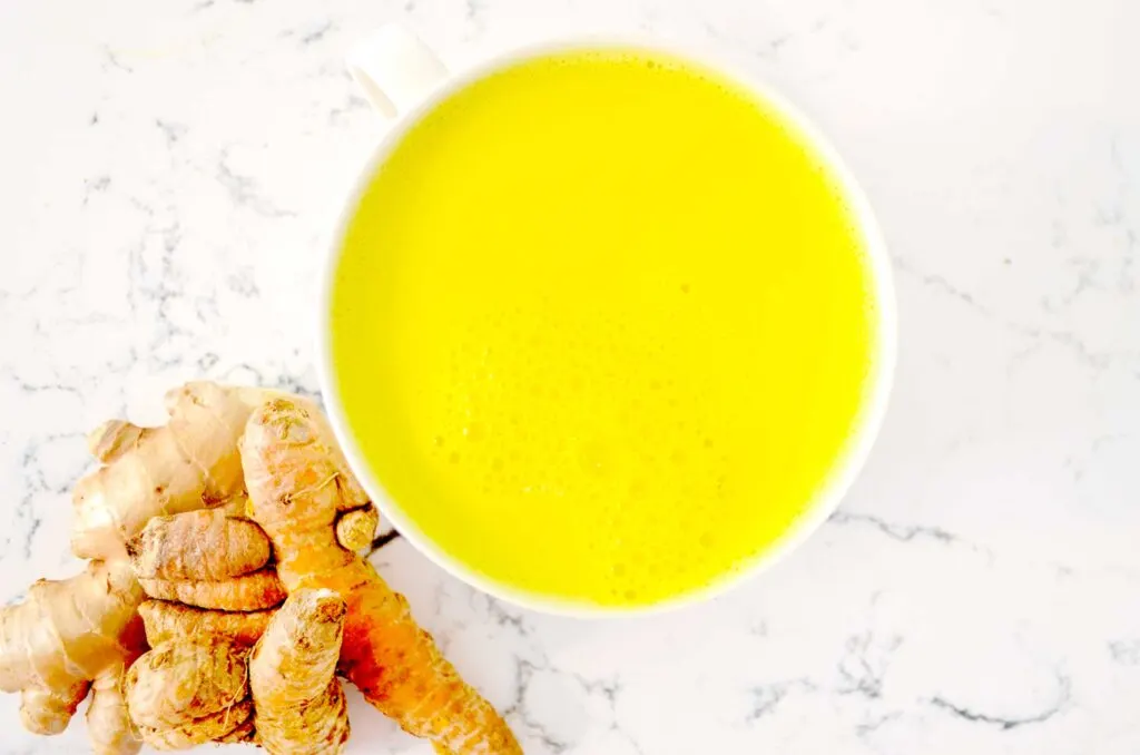 an overhead view of a bright yellow golden milk latte already made. There are bubble in the latte and the ingredients sit next to the mug, fresh ginger and turmeric root