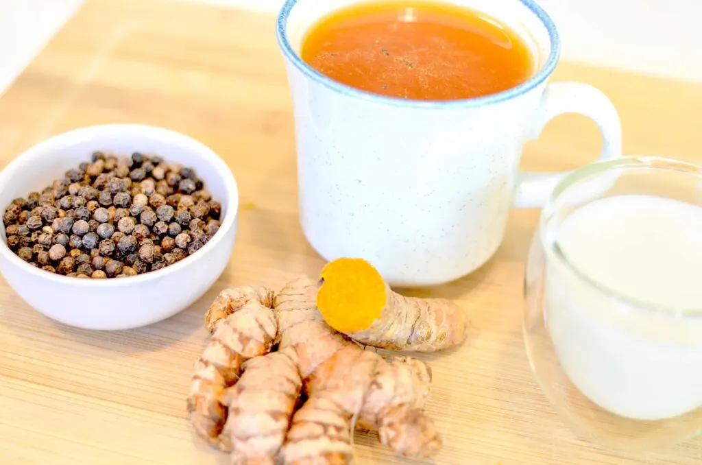 The ingredients you need to use in turmeric tea to absorb all the benefits. In the middle is a finalized cup of turmeric tea, with speckles of black pepper laying on top. In front of the mug, you have a pinch bowl with black peppercorns, the full turmeric root, and finally a mini double walled espresso mug filled with milk.