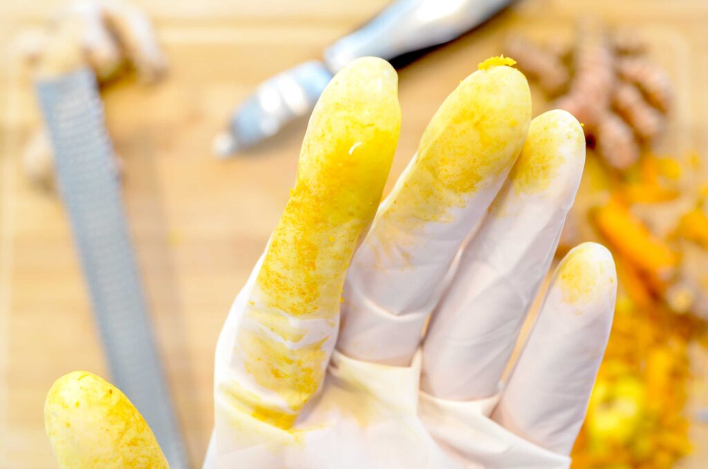 a close up of a gloved hand. What use to be a white glove has been stained to a golden yellow / orange from cutting and grating turmeric. Below the hand, slightly blurred is the aftermath of grating turmeric. On a cutting board, theres a grater, a peeler, a large orange pile, which is presumably the grated turmeric root.