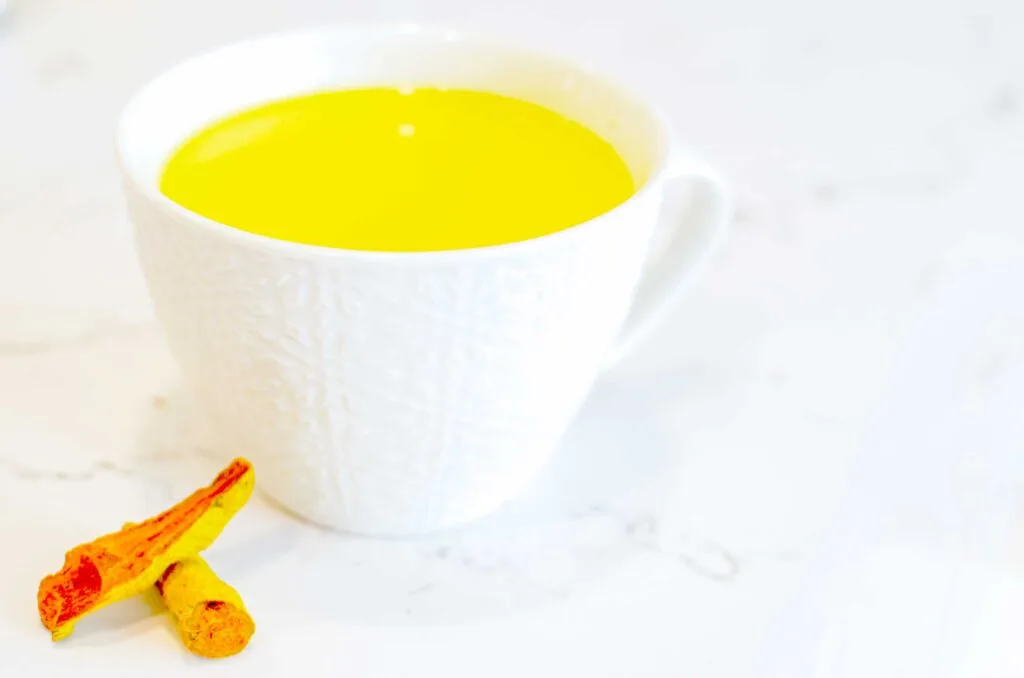 a simple photo of a marble counter top design with a white on white patterned embossed cappuccino mug. There's a bright yellow drink inside, a turmeric tea latte. The are two fresh stalks of vibrant orange turmeric leaning on each other and the mug