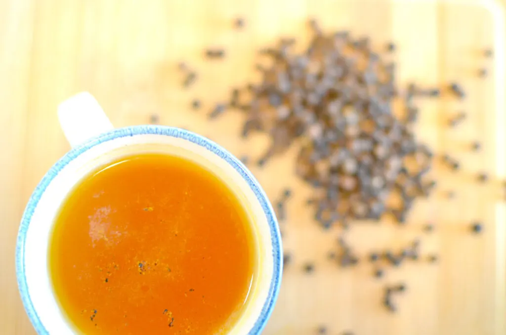 a top down view of an overexposed white speckled blue rimmed mug with a beautiful deep orange liquor inside. There are a few black speckles on top. Next to the mug is a pile of peppercorn.