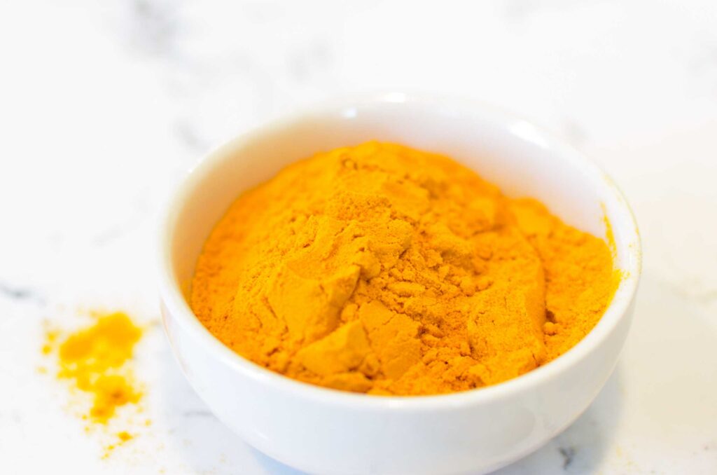 a close up of orange turmeric tea powder in a white punchbowl on a white and grey marbled counter top. A bit of the powder has spilled out onto the counter, staining the white to be yellow 