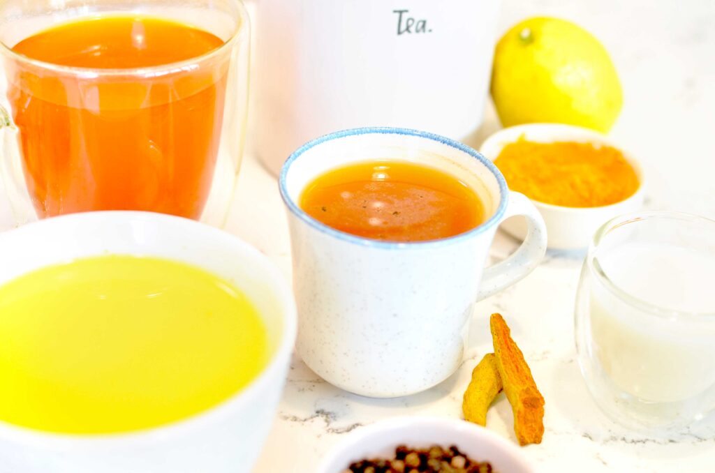 how to make turmeric tea, three options are shown with different ingredients to add. There are three mugs, a latte, a double walled bright orange mug, and a small mug with speckles of black pepper on top. Around the mugs are different ingredients, dried turmeric root, full peppercorns, turmeric powder, milk, and a lemon