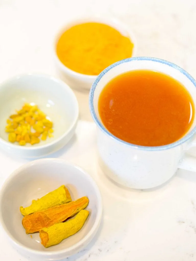 A vertical image showing all the types of turmeric you can use, and a small mug of deep orange turmeric tea in the middle. Each turmeric type is in a pinch bowl. There's a dried turmeric root, that looks like dried out carrots. Then there's dried turmeric that's been turned to pieces, and finally there's turmeric powder