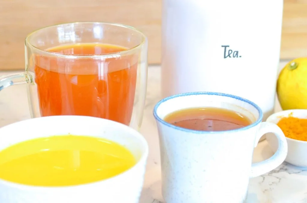 a close up of all different style and shaped mugs with different drinks inside. There's also a white jar behind them all that says: tea.
The three mugs with tea range in colours from a bright yellow, to a deep orange, and finally an orange brown. It's very autumn vibes. The mug in the front is short and wide, a cappuccino mug, behind it is a taller double walled mug, and finally a white speckled espresso mug with a blue rim sits in between both. 