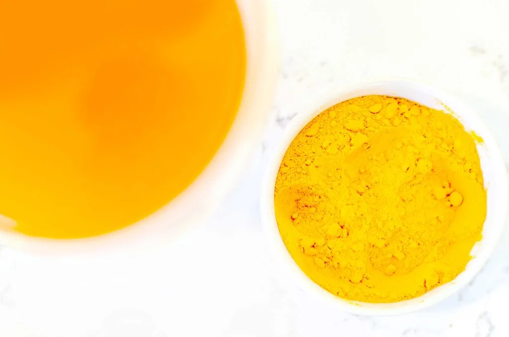 an overhead photo showing two circular kitchen ware items: a mug, and a pinch bowl. The mug has a deep orange brew, but inside the punch bowl is a heap of slightly lighter orange turmeric powder