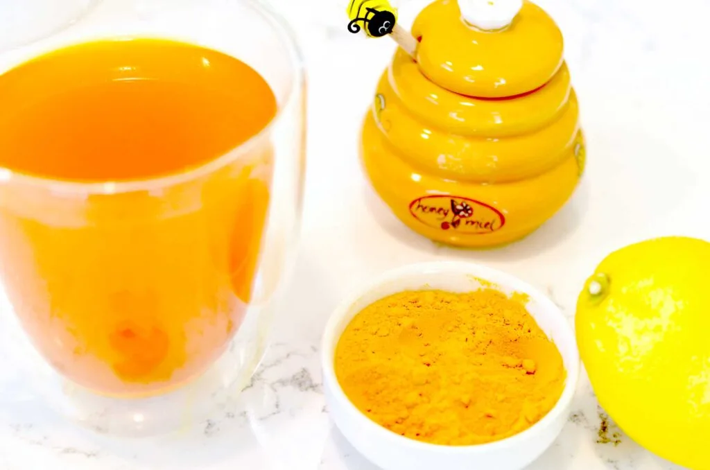 The ingredients you need to make a immune boosting healthy tea. There's a double walled mug with a beautiful orange tea inside. Next to it are the three ingredients used to make it: a jar of honey, turmeric powder, and a full lemon