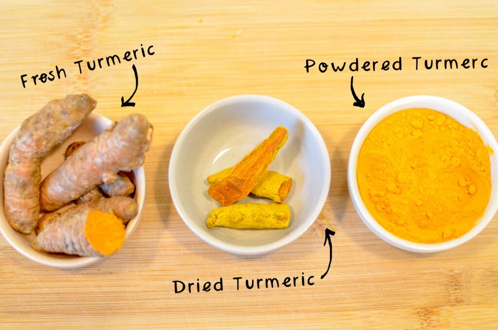 three pinch bowls filled with different stages of turmeric, plus arrows with text that describes each one. The first one is a fresh turmeric root, they look like thick brown misshappen skinnier sweet potatoes, with a bright orange inside. In the middle are shaved and dried pieces of the turmeric root, they still have an orange inside, but the outside is more yellow instead of brown, and they've shrunken. Finally, you have powdered turmeric, which is a bright orange powder