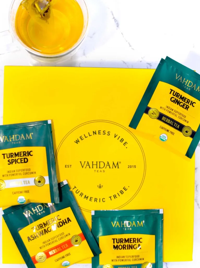 An overhead photo of a box of Vahdam Teas turmeric tea tribe, a simple golden yellow box with the logo in a vintage circle in the middle. There are four individual packaged tea bags around, showcasing what's in the box: turmeric spice, turmeric ginger, turmeric ashwadandha, and turmeric moringa. There's also a double walled mug sitting above it showcasing the beautiful steeped colour of these teas. 