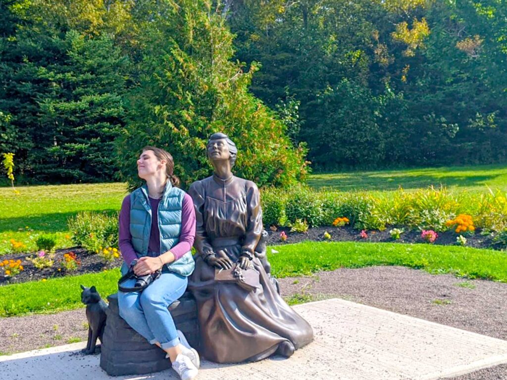 lucy maud-montgomery park statue. The bronze statue is in a long sleeve dress with a high neck and flowing down the floor, from the early 1900s. In her hands, is an opened, face down book. Her heda is titled up to towards the sun. Her hair is in a loosely fitted bun. She sits on a bench, with room next to her where a real woman sits in a similar pose, taking in the warm Prince Edward Island sun. Instead of a book on her lap, it's a DSLR camera. She's wearing clothes from the 21st century, a long sleeve shirt, a green puffer vest, with loose fitting jeans. Just behind the bench, is a bronze statue of a cat. Around the women, is a garden with flowers, grass and trees in the  background. 