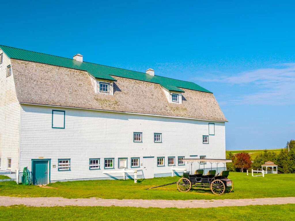 silver bush prince edward island. A giatn white painted barn takes up 2/3 of the photo. The tip top of the roof is painted in the classic Green Gables green. In front of the barn is an empty three row black horse drawn carriage. 

Next to the barn in the distance, looks like the tiniest gazebo, putting the size of the barn into perspective. 