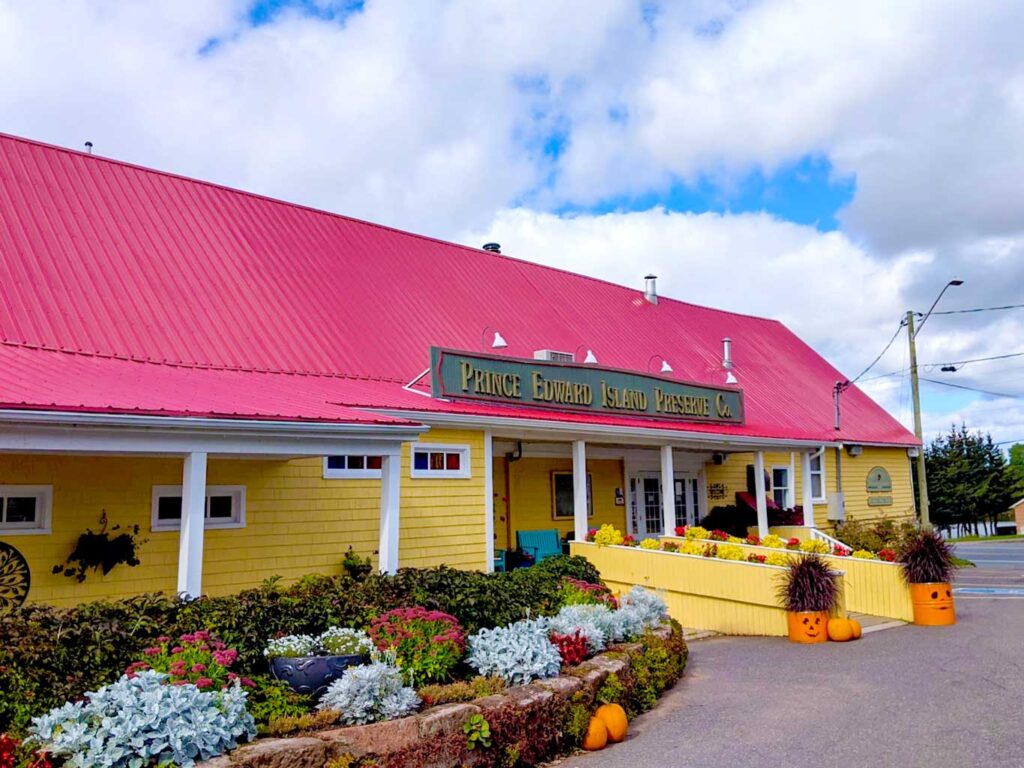 pei preserve company perfect place to shop for souvenirs and for tea. 

The image shows the outside of the building. A beautiful bright red roof, with yellow shingles siding the buidlings. There are a few small windows, and a ramp leading to the entrance. On the lowest part of the roof is a raised sign that says:  Prince Edward Island Preserve Co.

In front of the buildings is the start of the beautiful garden, with layers of bushes in the back and pink and light green flowers in the front. 