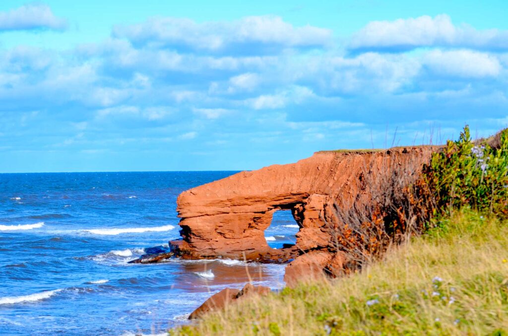 mackenzies brook sea arch prince edward island photographed in 2021. A large red sea wall juts out from the side of the cliffs edge, there's a small hole, creating an archway to walkthrough. 

The photograph was taken at high tide so the water is crashing in to the archway and the sea cliffs. 