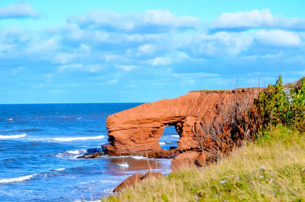 mackenzies brook sea arch prince edward island photographed in 2021. A large red sea wall juts out from the side of the cliffs edge, there's a small hole, creating an archway to walkthrough. The photograph was taken at high tide so the water is crashing in to the archway and the sea cliffs.